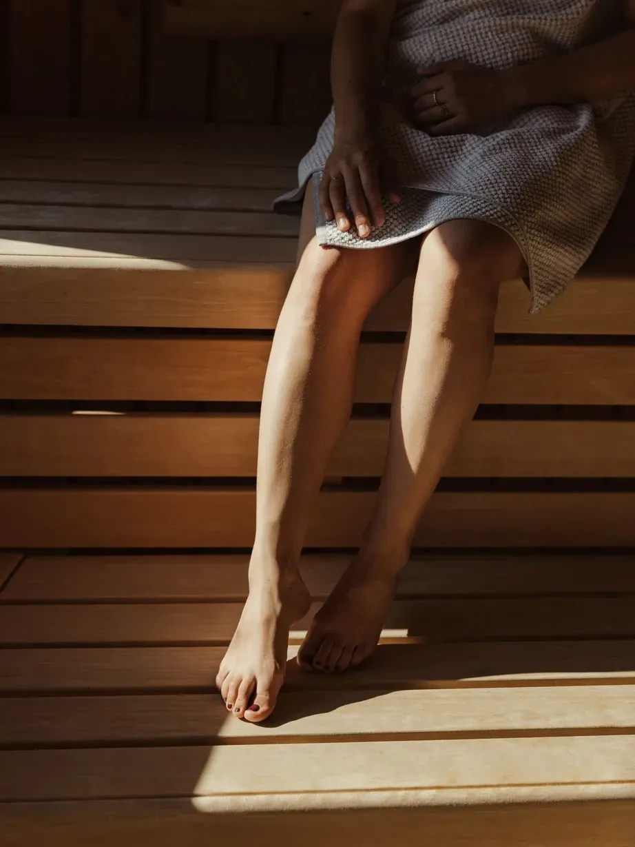 Una persona seduta su una panca di legno in sauna/spa, gambe nude e piedi, indossa un vestito chiaro a fantasia; la luce crea ombre sul pavimento.