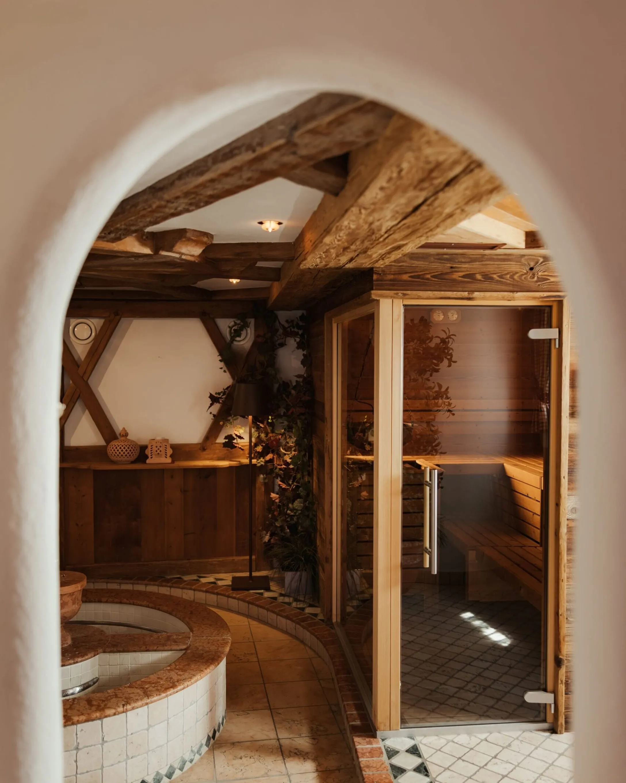 Interno spa dell'Hotel Gran Ander, Badia: arco in primo piano, vasca rotonda in mosaico, pavimenti a piastrelle, travi in legno e sauna in vetro a destra, luci calde.