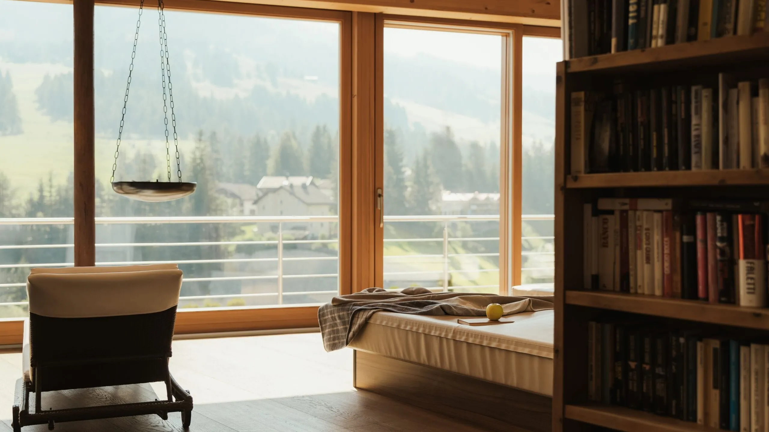 Interno accogliente al Hotel Gran Ander, Badia: sedia vicino a grandi finestre con vista sulle Dolomiti, letto e una libreria piena di libri.