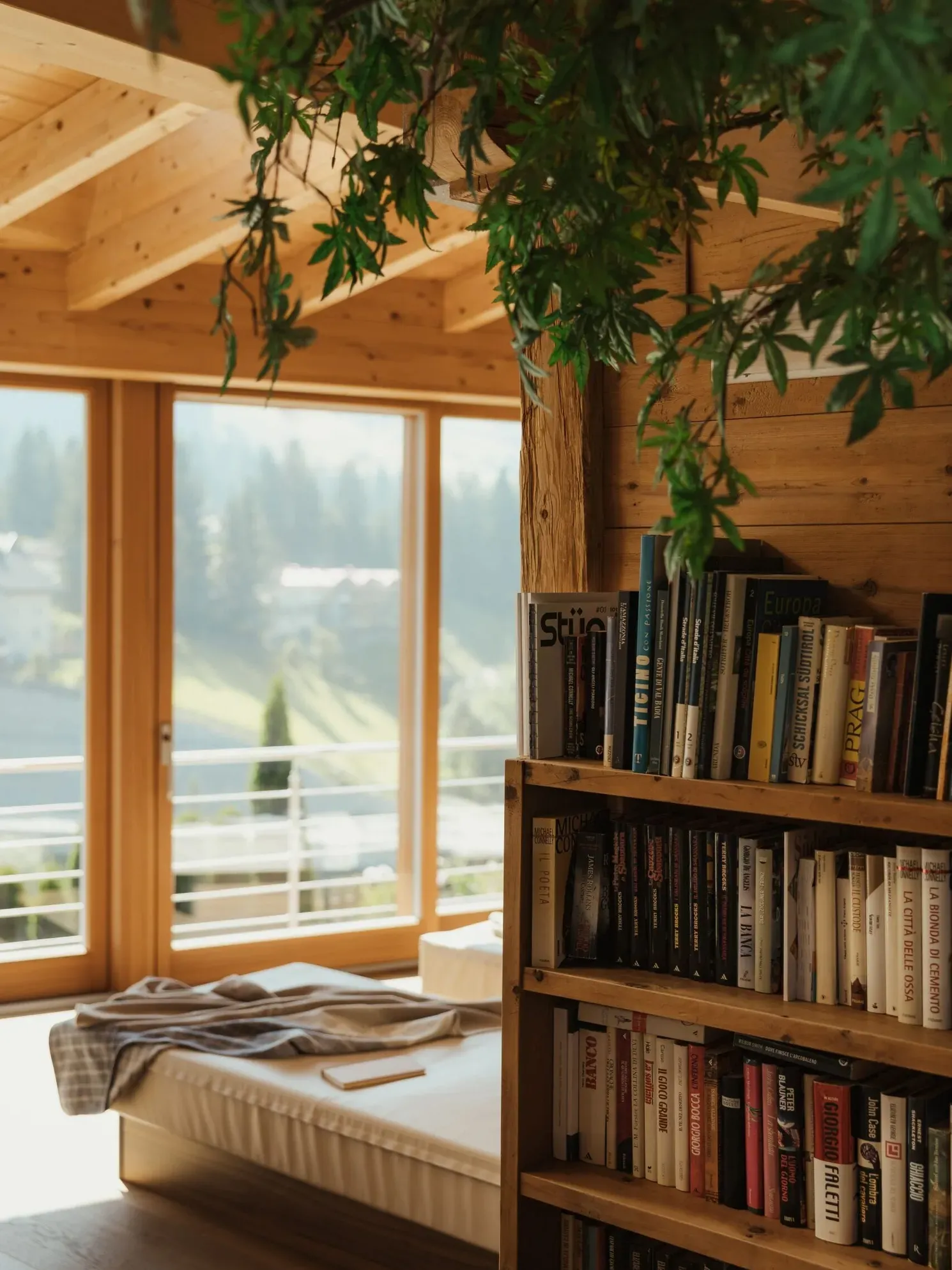 Interno in stile baita: legno a pareti e soffitto, letto accanto a una grande finestra con vista sulle Dolomiti; scaffale di libri e piante pendenti.
