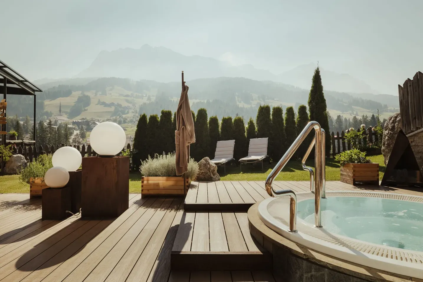 Alpine pool deck in Alta Badia with a circular pool, stainless rails, wooden decking, lounge chairs, spherical lamps, potted plants, and Dolomites.