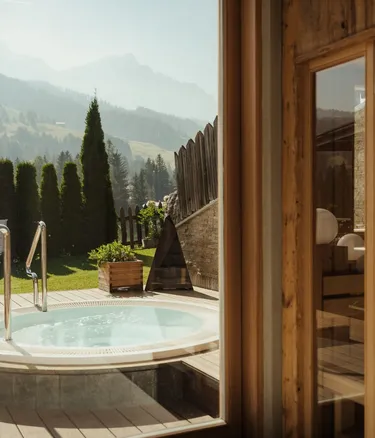 Jacuzzi rotonda su terrazza in legno, con giardino e Dolomiti sullo sfondo; vetri e legno dell'interno incorniciano la scena.