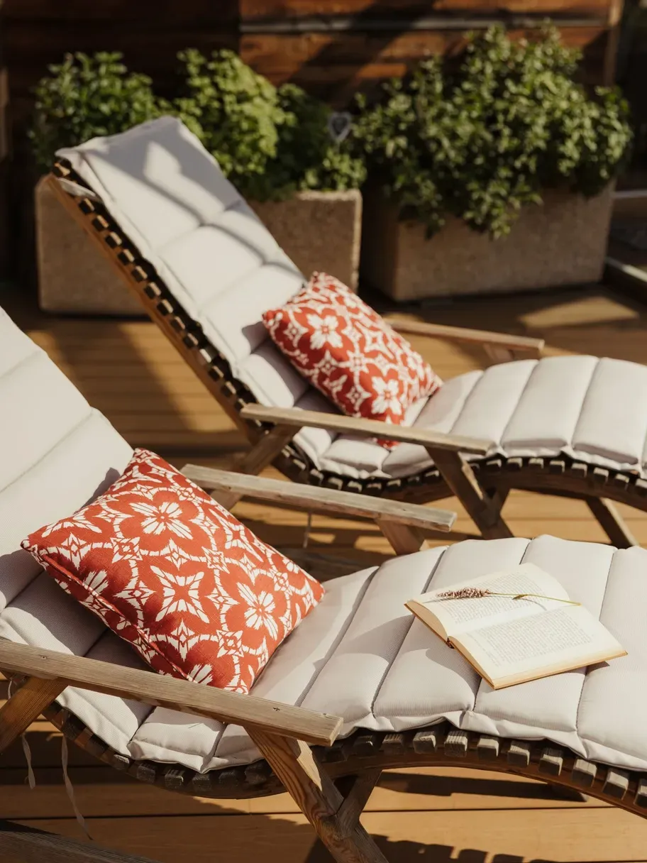 Outdoor wooden deck with cushioned lounge chairs, red patterned pillows, an open book, and potted green shrubs against a wooden wall.