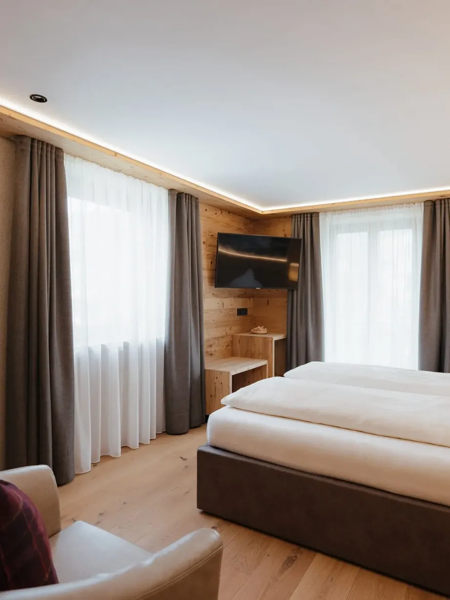 Cozy hotel room with light wood walls and floor, two beds, wall-mounted TV, seating area with plaid cushions, and large windows with sheer curtains.