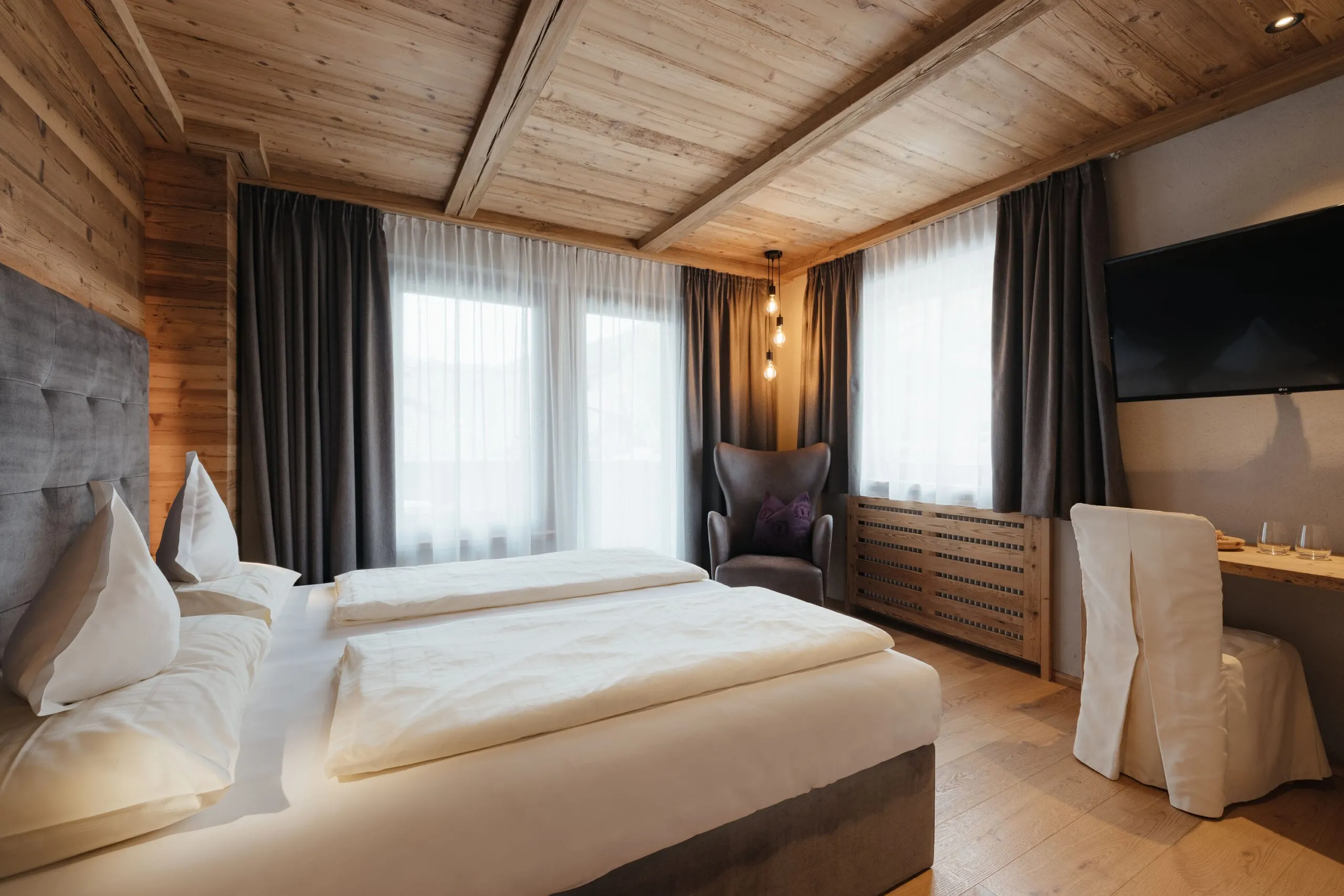 Hotel Gran Ander bedroom with timber ceiling and walls, a large bed in white linens, a gray headboard, a reading chair, desk, curtains, and a wall TV.