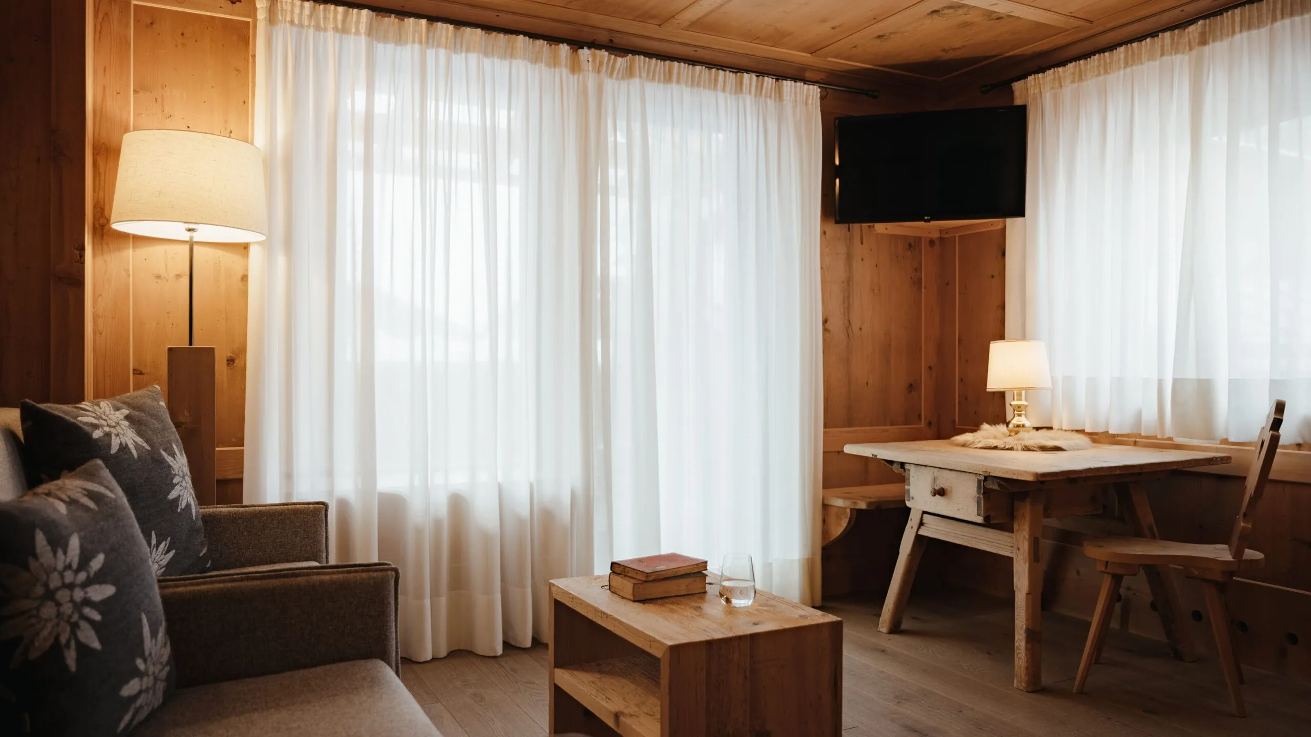 Interno in stile chalet di legno: divano con cuscini, tavolino, tende bianche filtranti la luce, tavolo da pranzo con sedie e TV a parete.