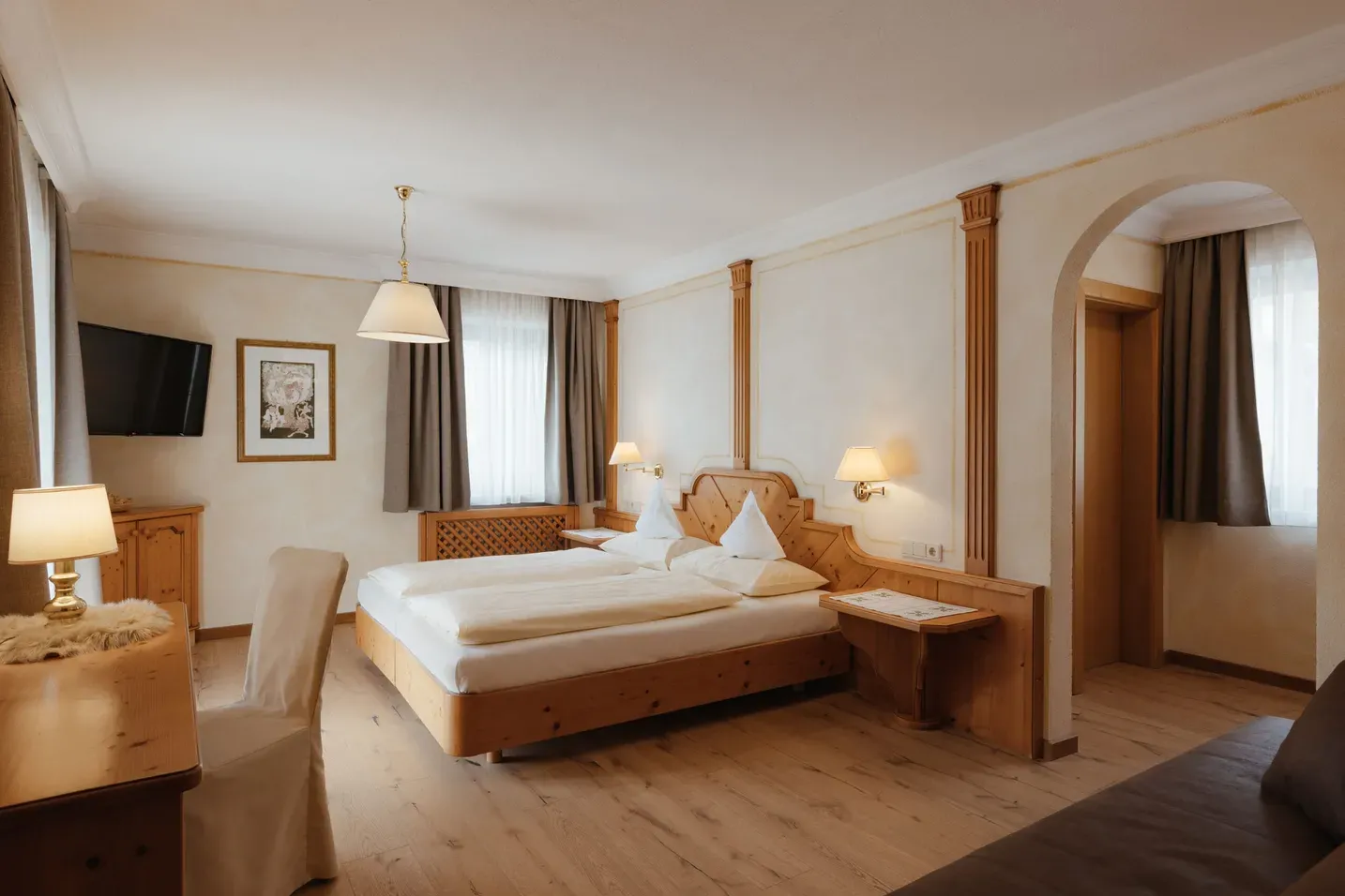 Helles Alpenhotelzimmer mit Holzmöbeln, großem Doppelbett, zwei Nachttischen, Schreibtisch, Wand-TV, Fenster mit Vorhängen und einer Bogenöffnung.