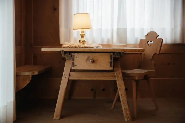 Rustikales Hotelzimmer mit massivem Holztisch, geschnitztem Stuhl und kleiner Lampe; warmes Licht fällt durch helle Vorhänge auf die Holzwand.