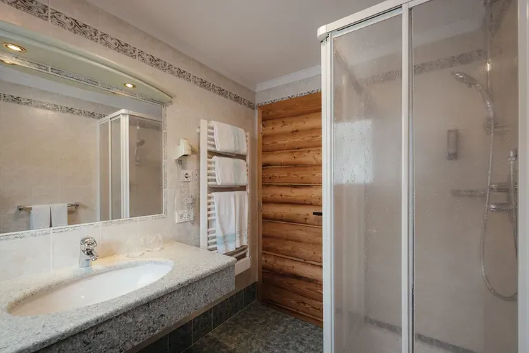 Helles Hotelbadezimmer mit Granit-Waschtisch, beleuchtetem Spiegel, Handtuchheizung und weißen Handtüchern, Holztür-Verkleidung und Glas-Duschabtrennung.