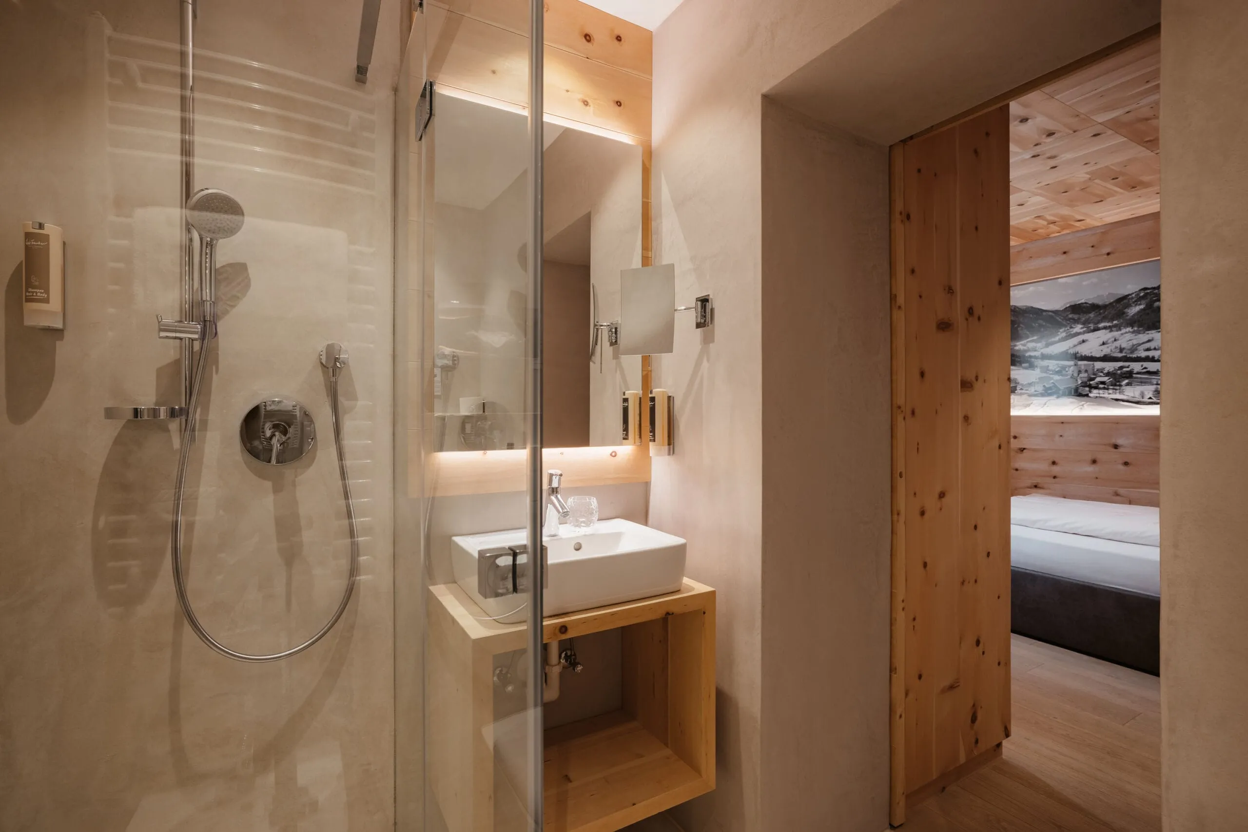 Modernes Badezimmer im Hotel Gran Ander mit Glas-Duschkabine, Waschbecken auf Holztisch, beleuchtetem Spiegel und Durchgang zum Schlafzimmer mit Holzverkleidung.