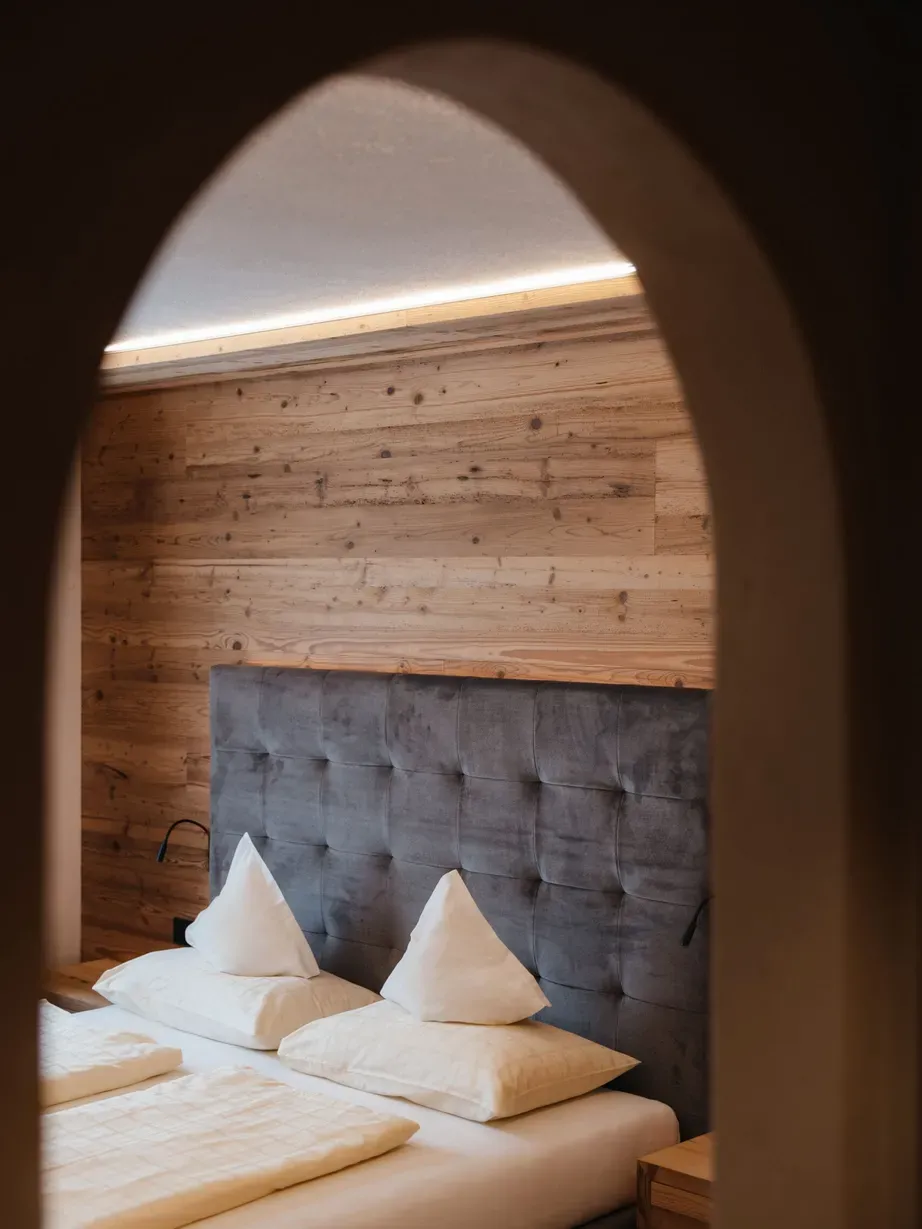 Hotel Gran Ander, Badia: Blick durch einen arkadenförmigen Türbogen auf ein Bett mit dunkelgrauem Kopfteil, Holzpaneelwand und weißen Kissen.