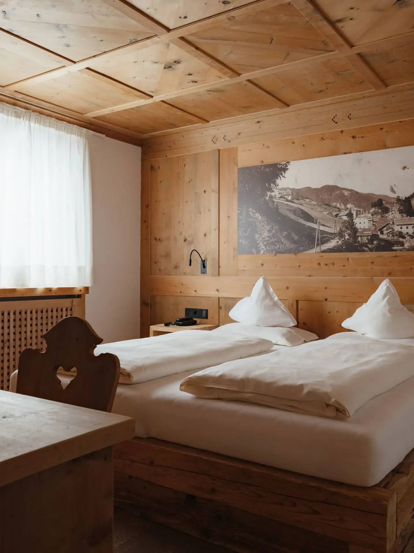 Holzverkleidetes Hotelzimmer mit Doppelbett, zwei weißen Kissen, Nachttischen, Wandbild einer Dolomitenlandschaft, Fenster mit weißem Vorhang und Holztisch mit Snack-Schale im Vordergrund.