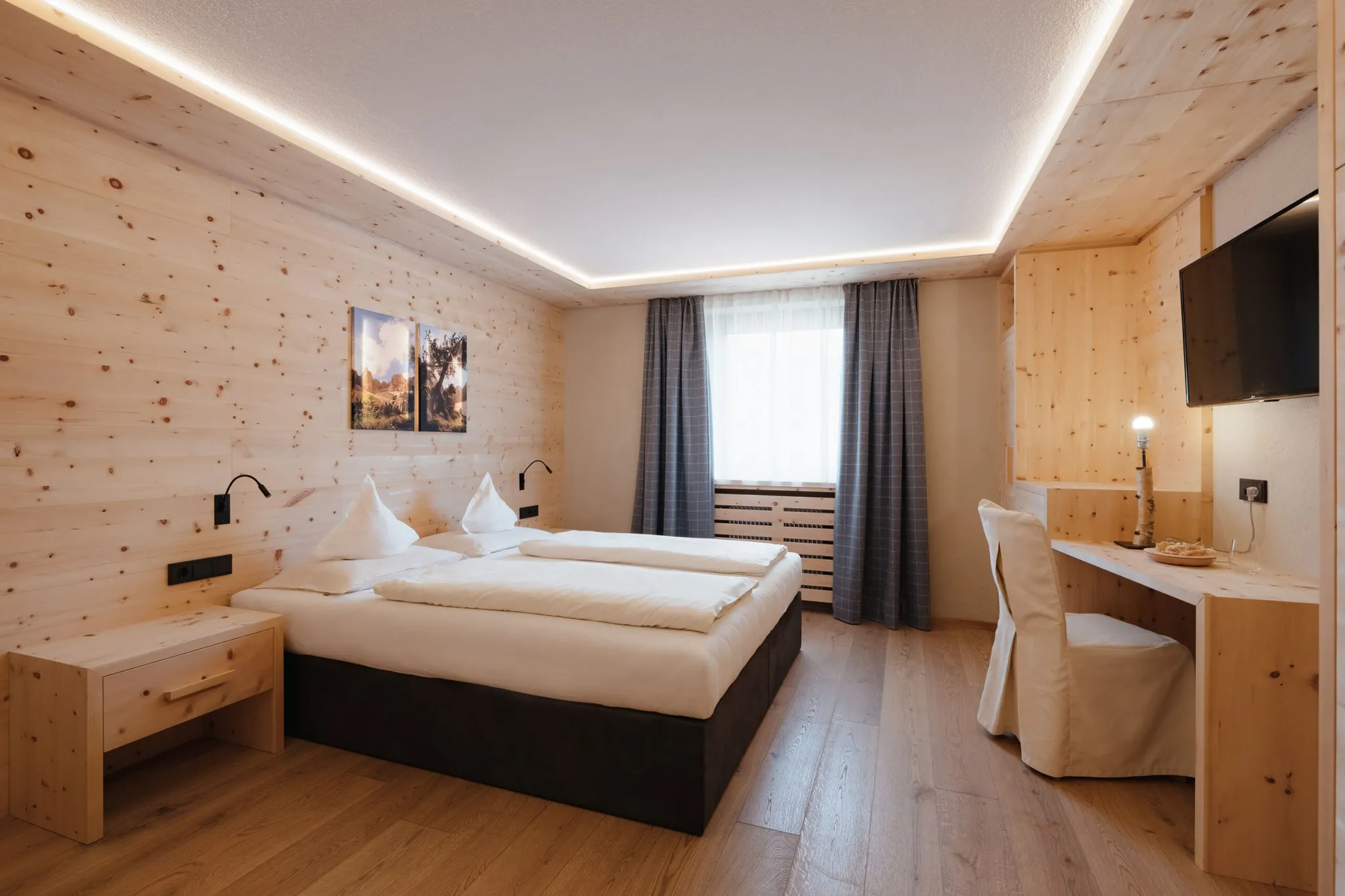 Helles Hotelzimmer mit Holzvertäfelung, zwei zusammengestellten Betten, weißer Bettwäsche, Nachttischen, Wandlampen, Schreibtisch, Stuhl, TV und Fenster mit Vorhängen.