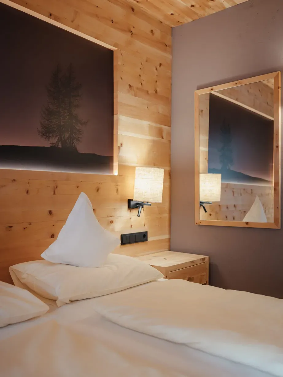 Rustikales Hotelzimmer mit Holzvertäfelung, Doppelbett mit weißen Kissen, Nachttisch, Wandlampen und Spiegel; Baum-Silhouette an der Wand.
