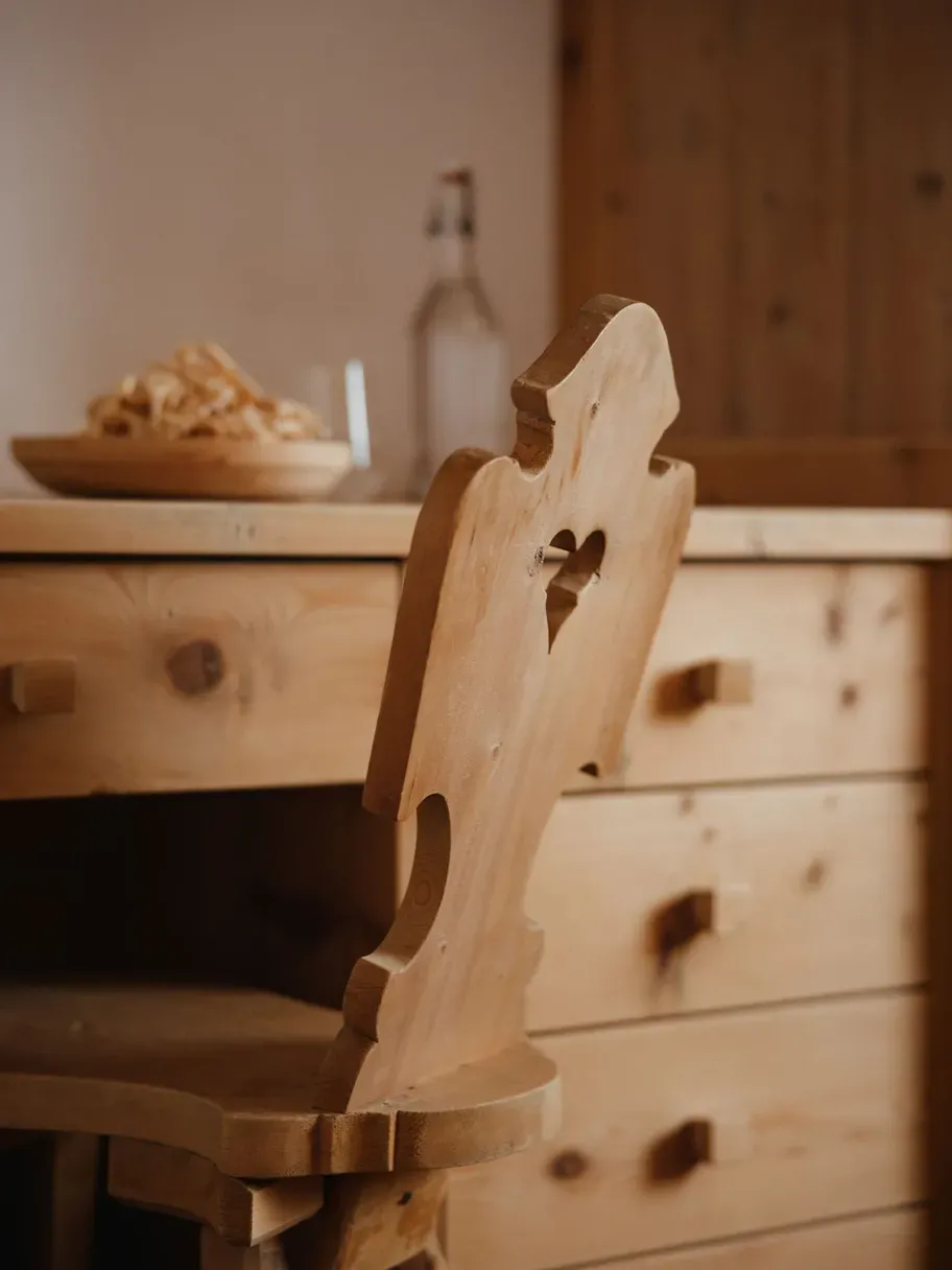 Close-up of a carved wooden chair back in a warm, rustic hotel interior, with a wooden dresser and a blurred plate of snacks in the background.