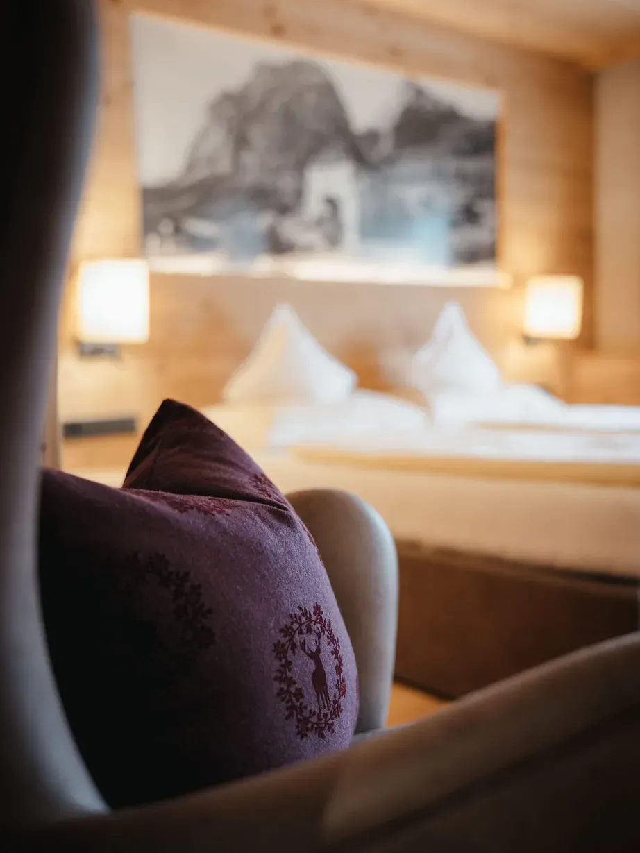 Cuscino viola ricamato in primo piano, letto bianco sullo sfondo, pareti di legno e luci calde in una camera d'albergo.