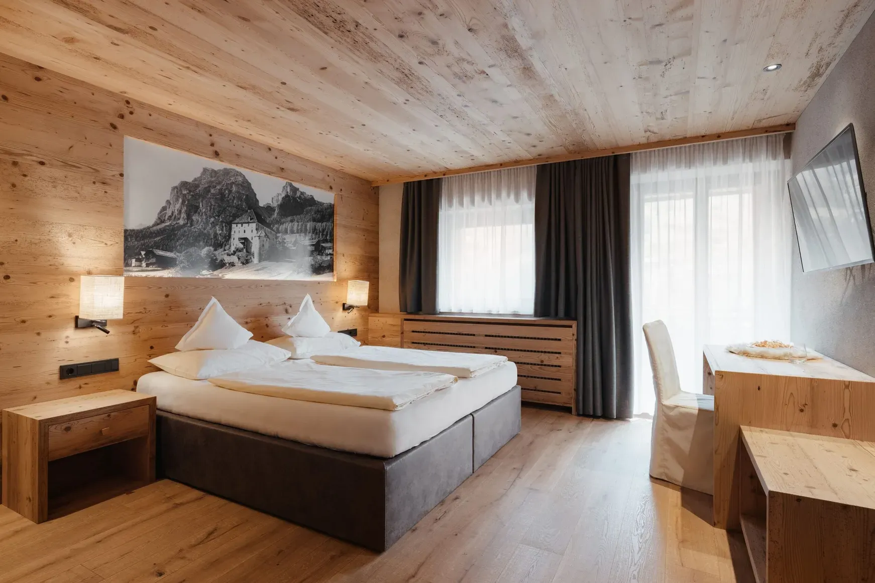 Hotelzimmer aus Holz: zwei Betten, Nachttische, Wandlampen, Fenster mit weißen Vorhängen, Schreibtisch und TV; Wandbild eines Bergschlosses.