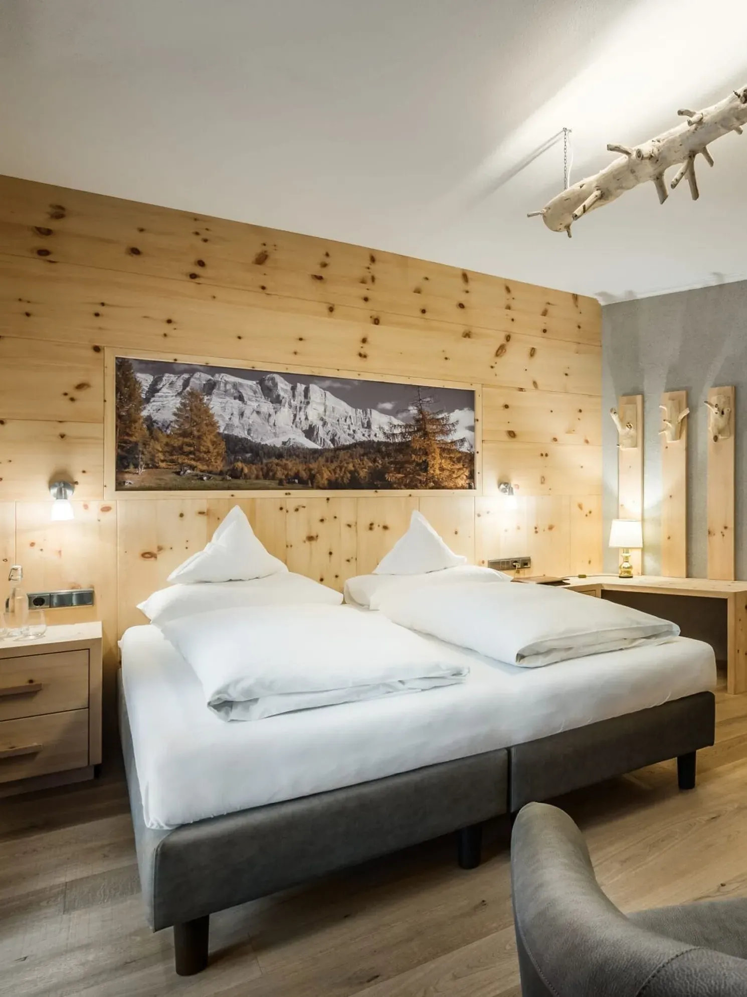 Hotel Gran Ander, Badia: Doppelbett vor Holzpaneelwand, zwei Nachttische, Schreibtisch, Garderobe, graues Sofa, Bergbild über dem Bett, Holzboden.