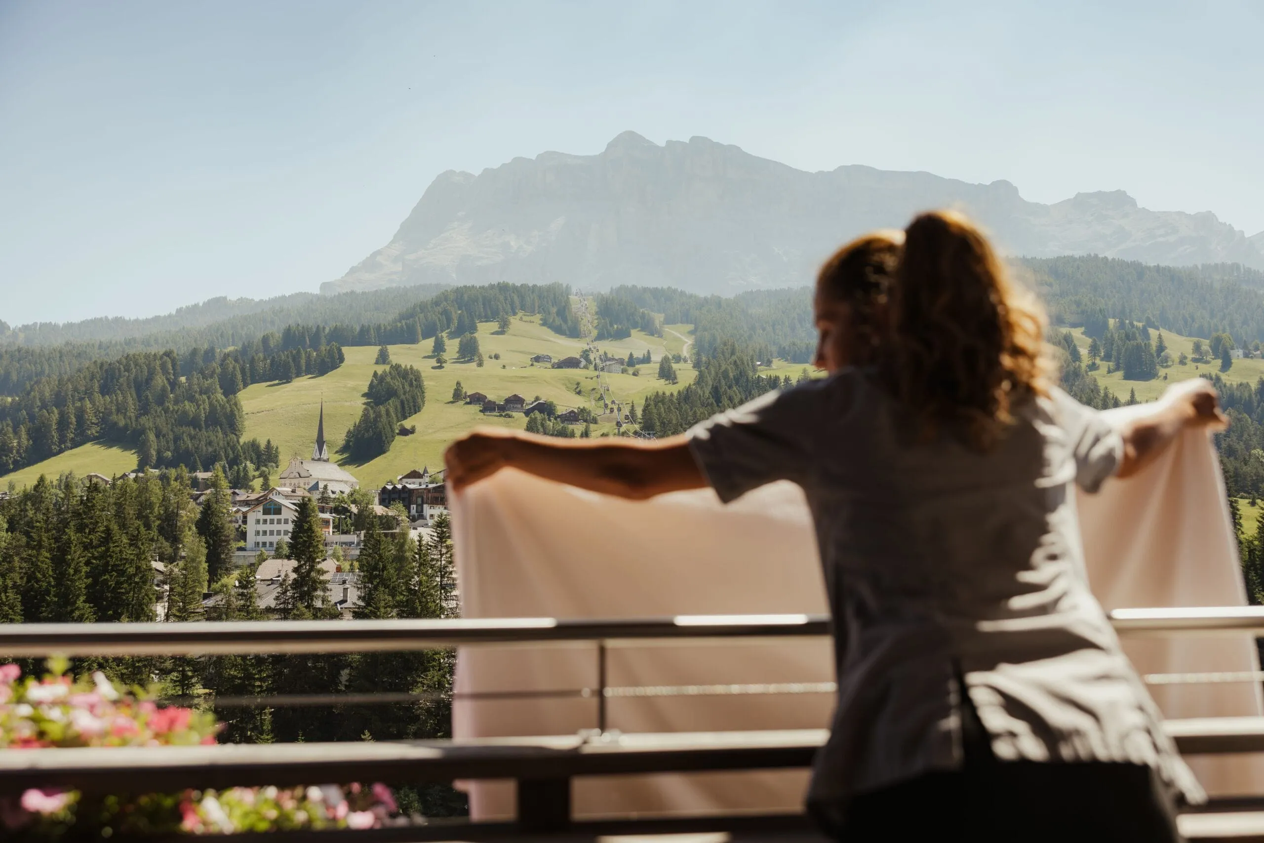 Person steht mit ausgestreckten Armen auf Balkon und blickt über Alta Badia in den Dolomiten: Dorf mit Kirchturm, Wälder und Bergkette im Hintergrund.