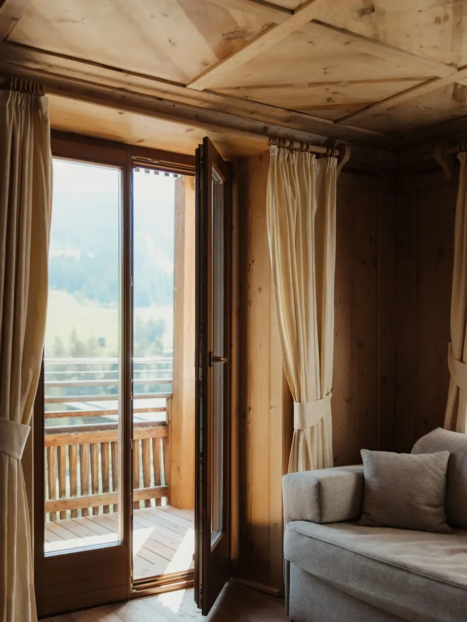 Wohnzimmer mit Holzverkleidung und Holzdecke, offene Balkontüren zum Holzbalkon, graues Sofa und beige Vorhänge; Berglandschaft im Blick.