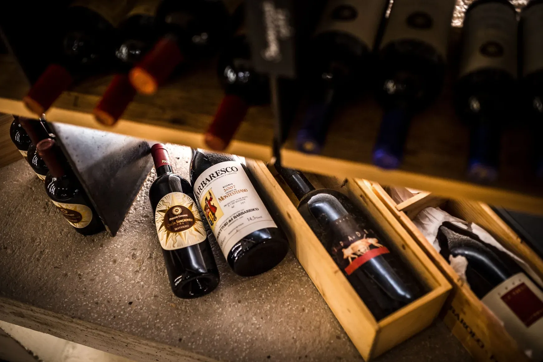 Bottiglie di vino adagiate su scaffali e in una cassa di legno, in una cantina, illuminate da una luce calda che mette in risalto le etichette.