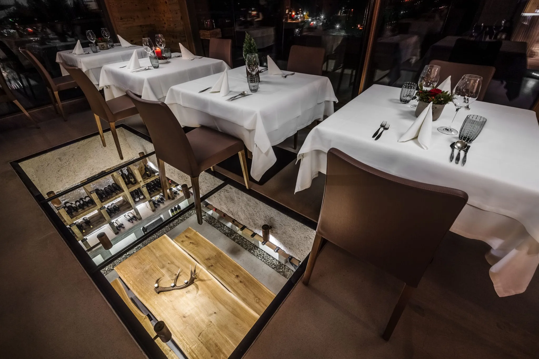 Ristorante interno elegante con tavoli apparecchiati e tovaglie bianche; pavimento a vetro mostra la cantina di vini sottostante.