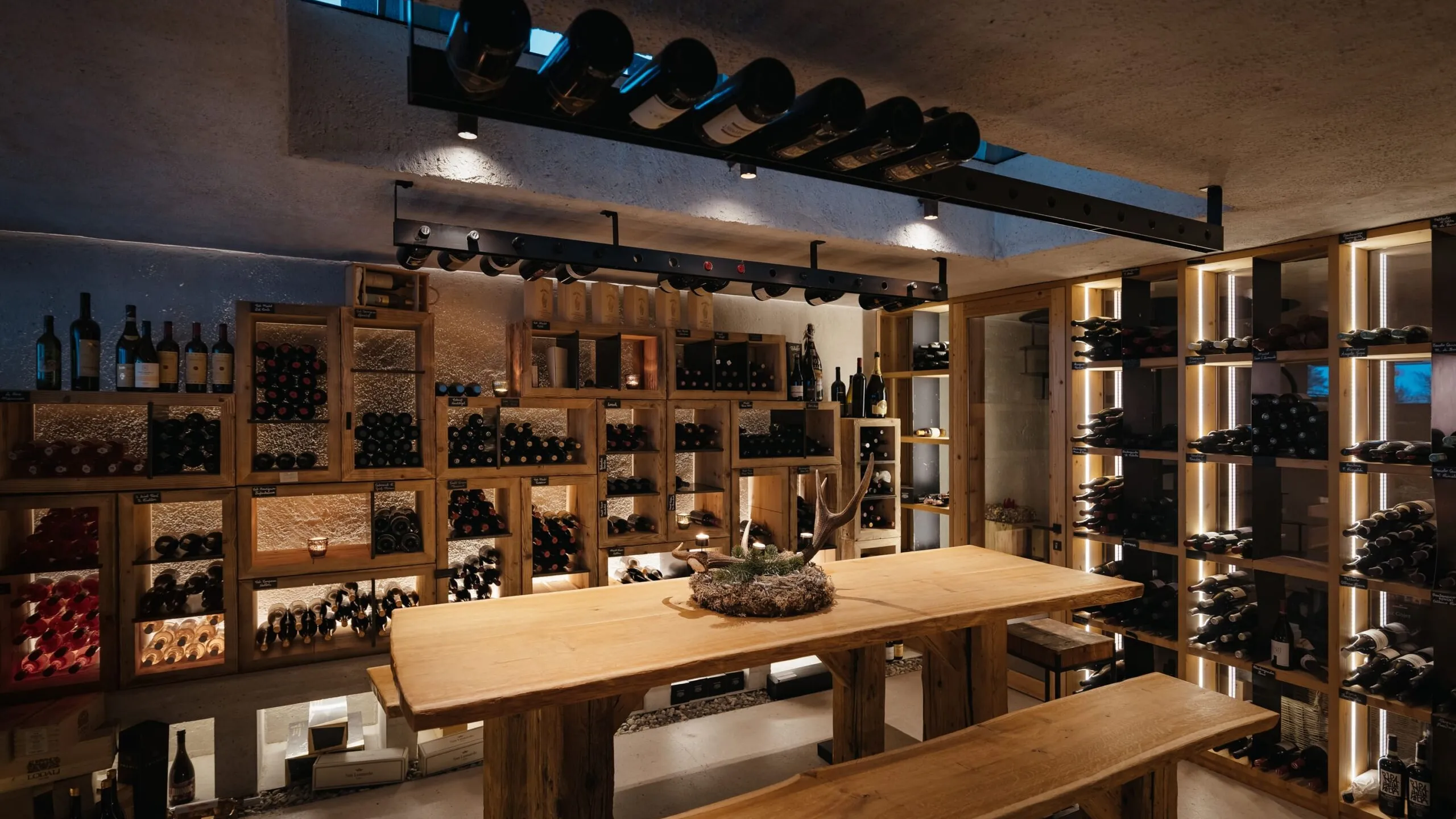 Enoteca in legno con scaffali a muro pieni di bottiglie, tavolo lungo al centro con centrotavola; atmosfera calda e accogliente.