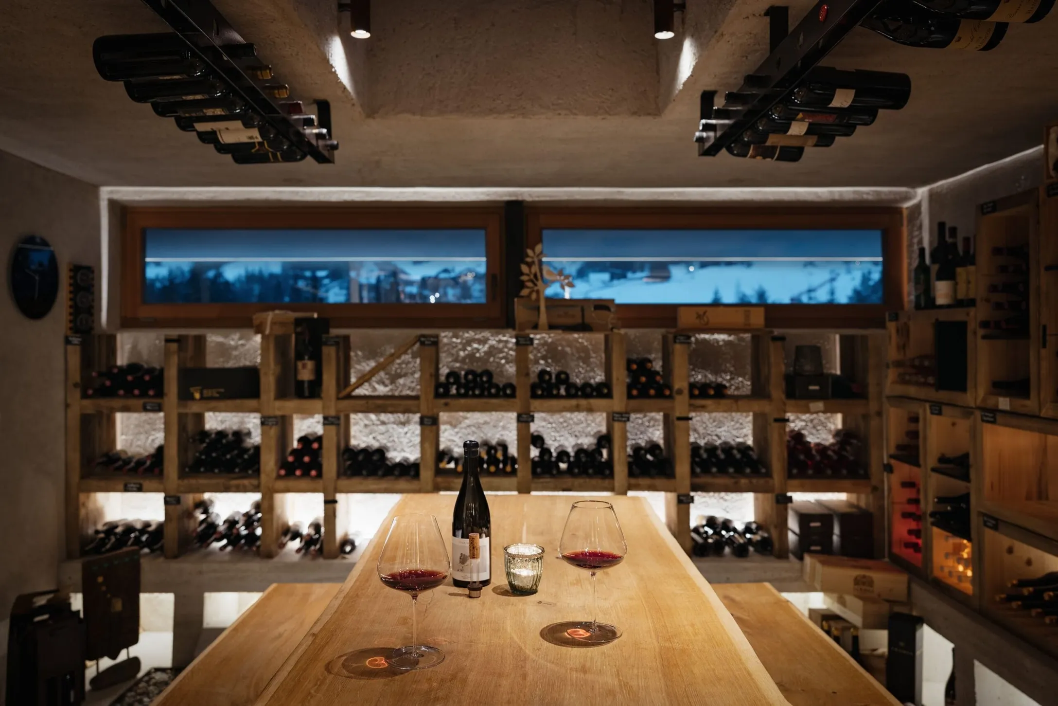 Cozy wine cellar with wooden bottle shelves, a central wooden table, a bottle and two red wine glasses, a small candle, and a snowy window in back.
