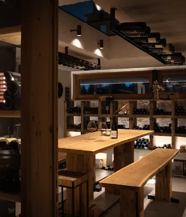 Interno rustico dell'Hotel Gran Ander, Badia: tavolo e panche in legno, scaffali con bottiglie, cantina/vineria illuminata da luci calde.