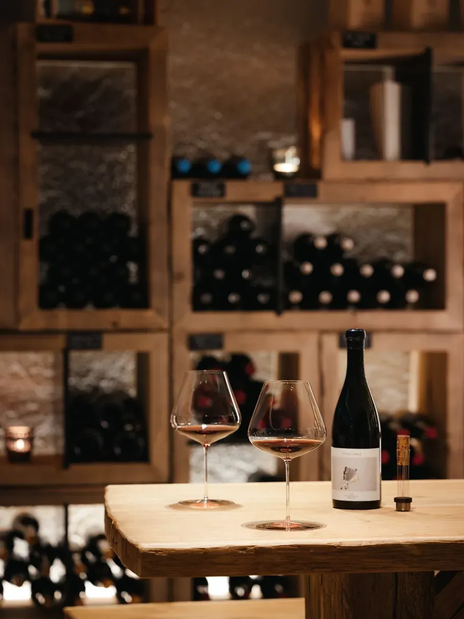 Two glasses of red wine and a bottle sit on a rustic wooden table, with warm shelves of bottles in the blurred background.