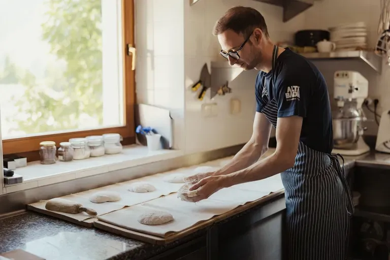Person in a striped apron and glasses kneads dough on a flour-dusted counter in a bright kitchen; jars line the windowsill beside a mixer in the background.