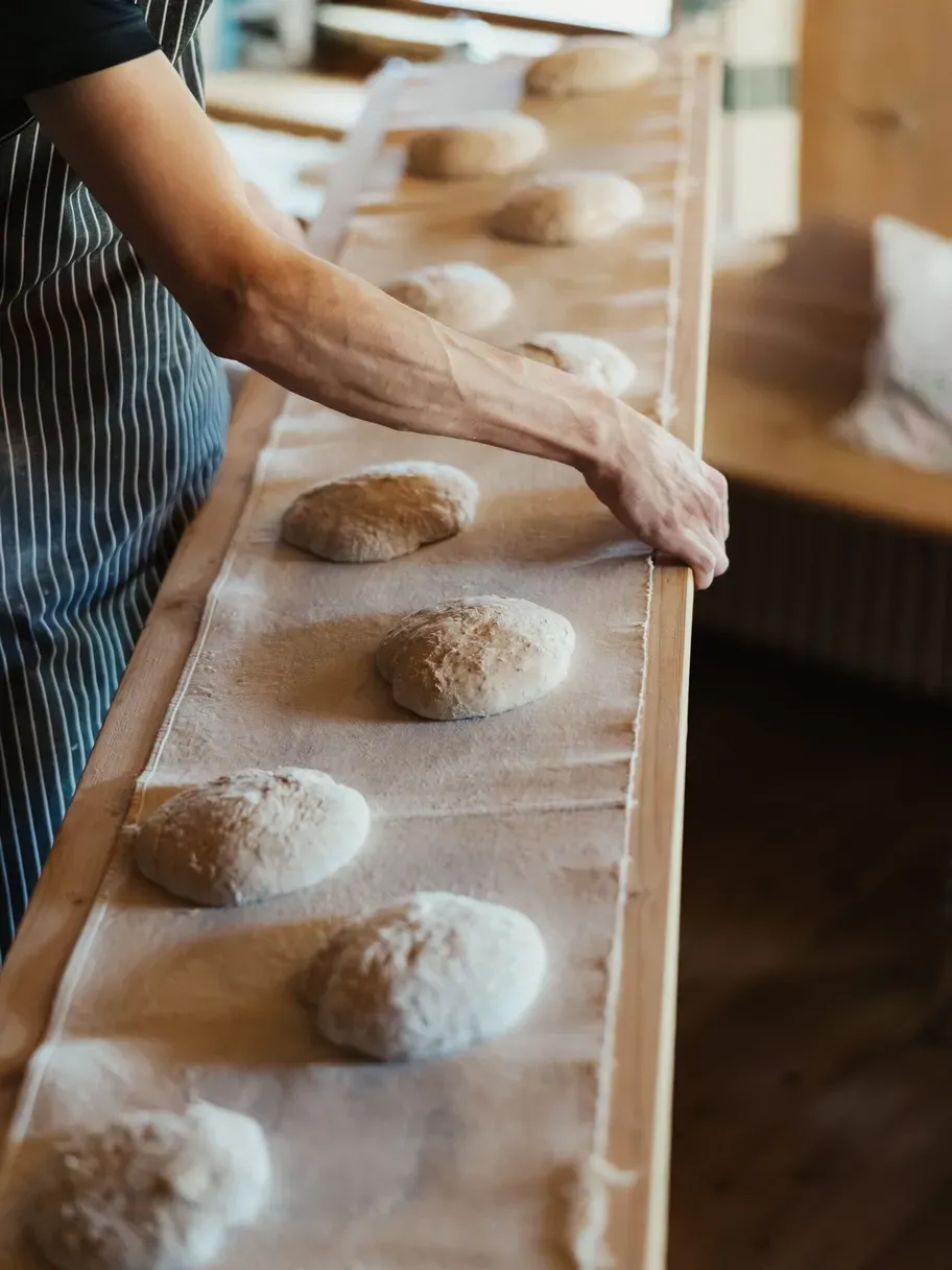 Una persona con grembiule a righe lavora su una tavola di legno, formando panetti di impasto infarinato allineati.