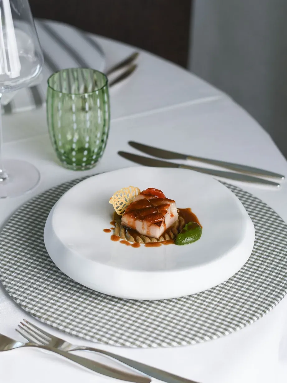 Piatto gourmet: pesce glassato su base di rösti, salsa marrone, cialda croccante e salsa verde, su piatto bianco.