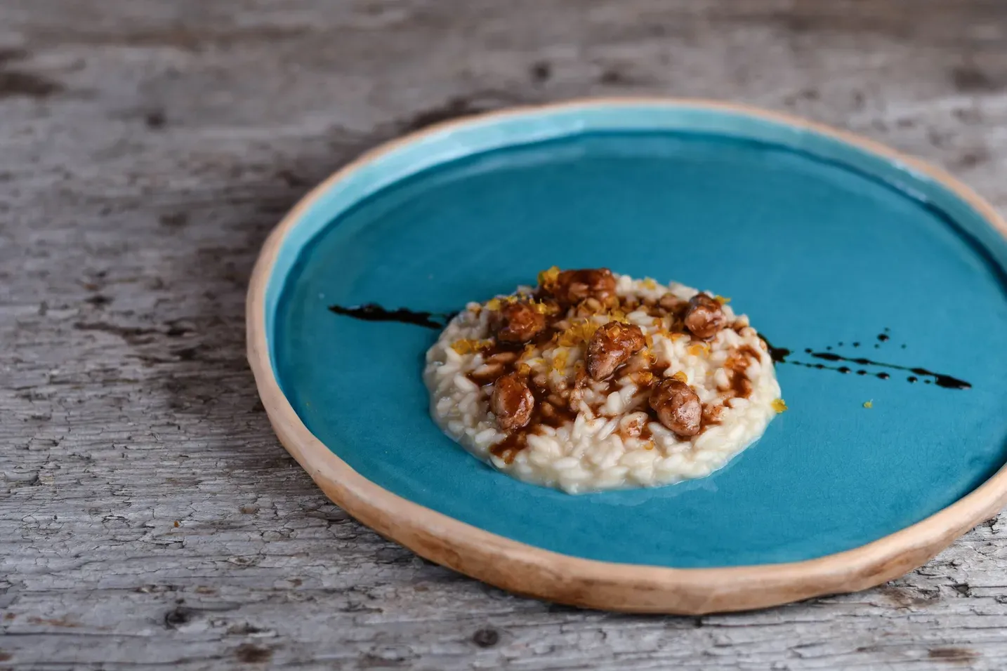 Risotto cremoso su piatto turchese, guarnito con croccante topping dorato e una riga di salsa scura, su tavola di legno.