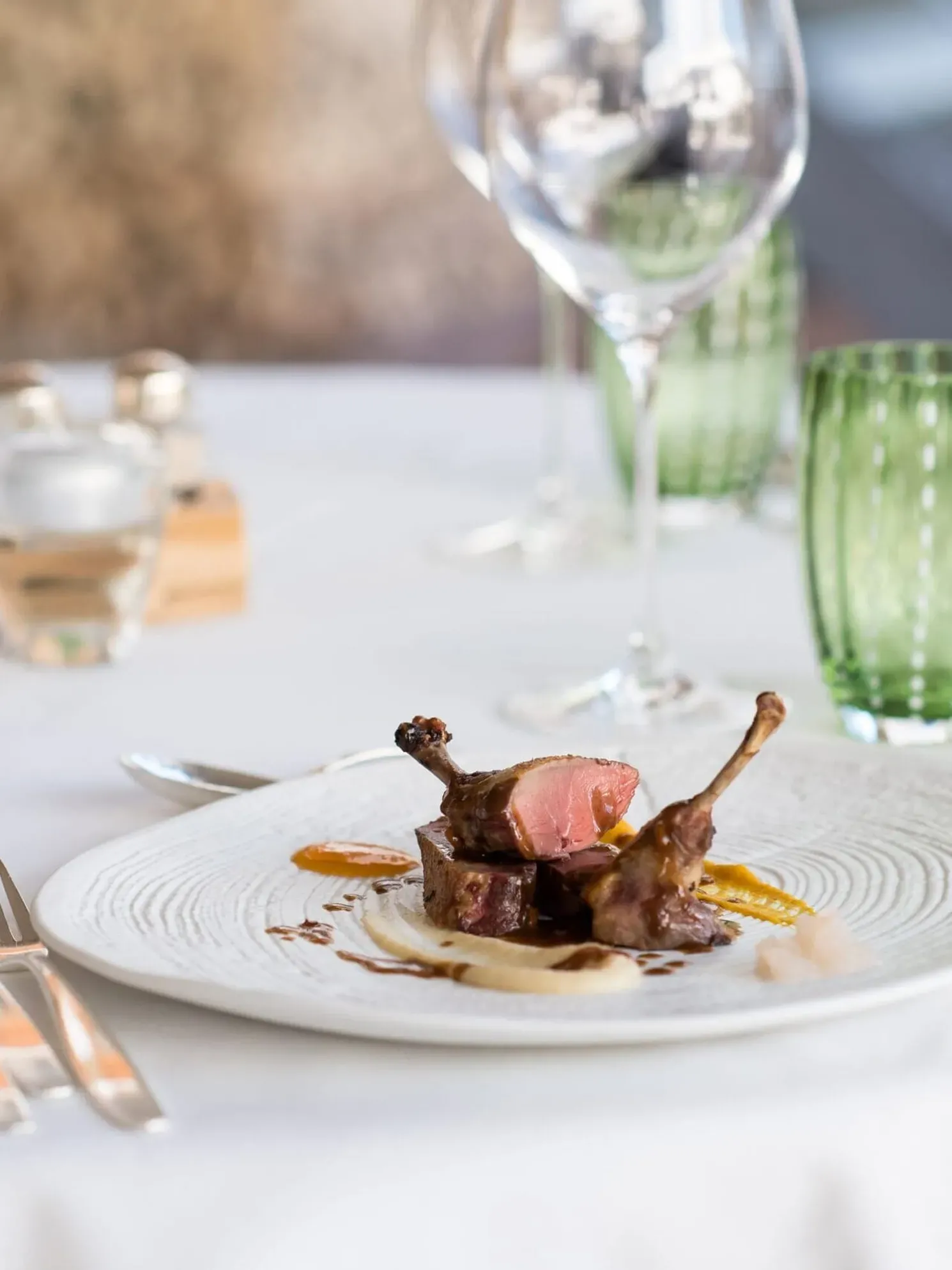 Piatto di carne con ossa, salsa marrone e purè, su piatto bianco, con posate ai lati e bicchieri verdi sul tavolo imbandito.