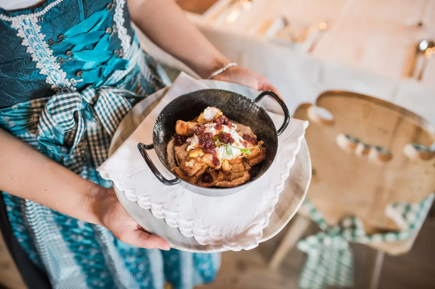 Una persona in abito tradizionale azzurro tiene un piccolo tegame nero con dolce fritto guarnito di salsa di frutti rossi, su piatto bianco con tovagliolo.