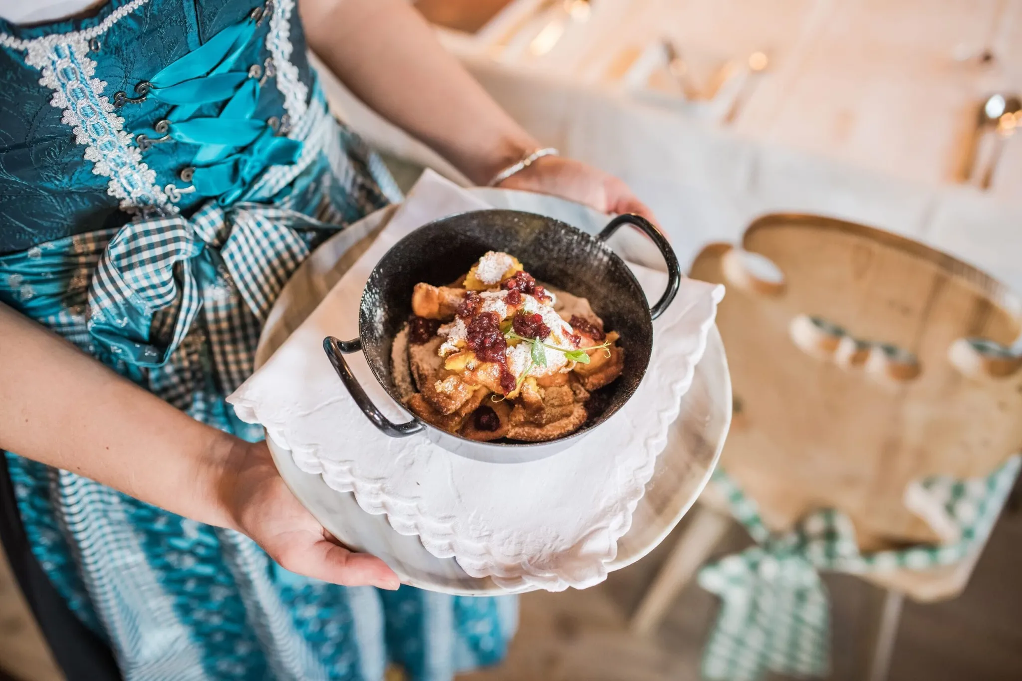 Una persona in abito tradizionale azzurro tiene un piccolo tegame nero con dolce fritto guarnito di salsa di frutti rossi, su piatto bianco con tovagliolo.