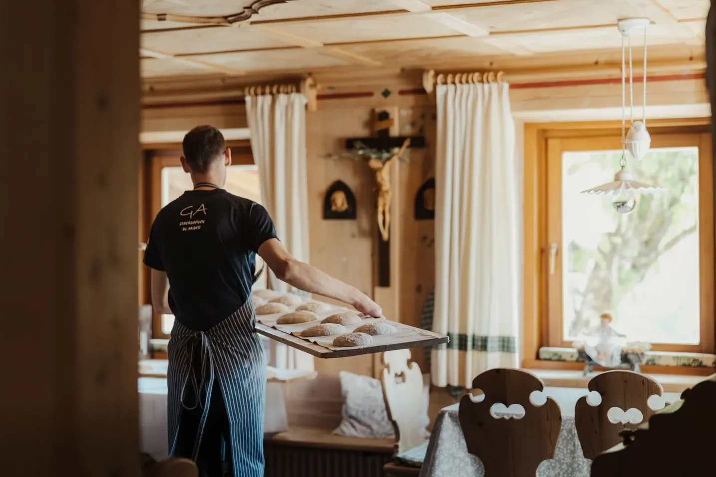 Persona di spalle in grembiule a righe tiene un vassoio con piccole pagnotte in una cucina rustica dell'Hotel Gran Ander, Badia, Dolomiti, con soffitto in legno e finestra luminosa.