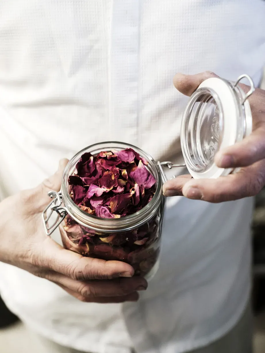 Una persona tiene un barattolo di vetro con coperchio aperto, pieno di petali di rosa secchi di colore viola-rosa.