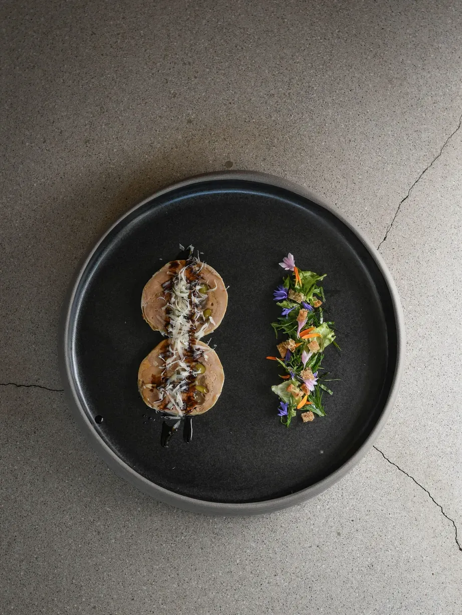 Piatto nero rotondo con due mini panini farciti e scaglie di formaggio, accanto a un'insalata colorata con fiori commestibili, su tavola di cemento.