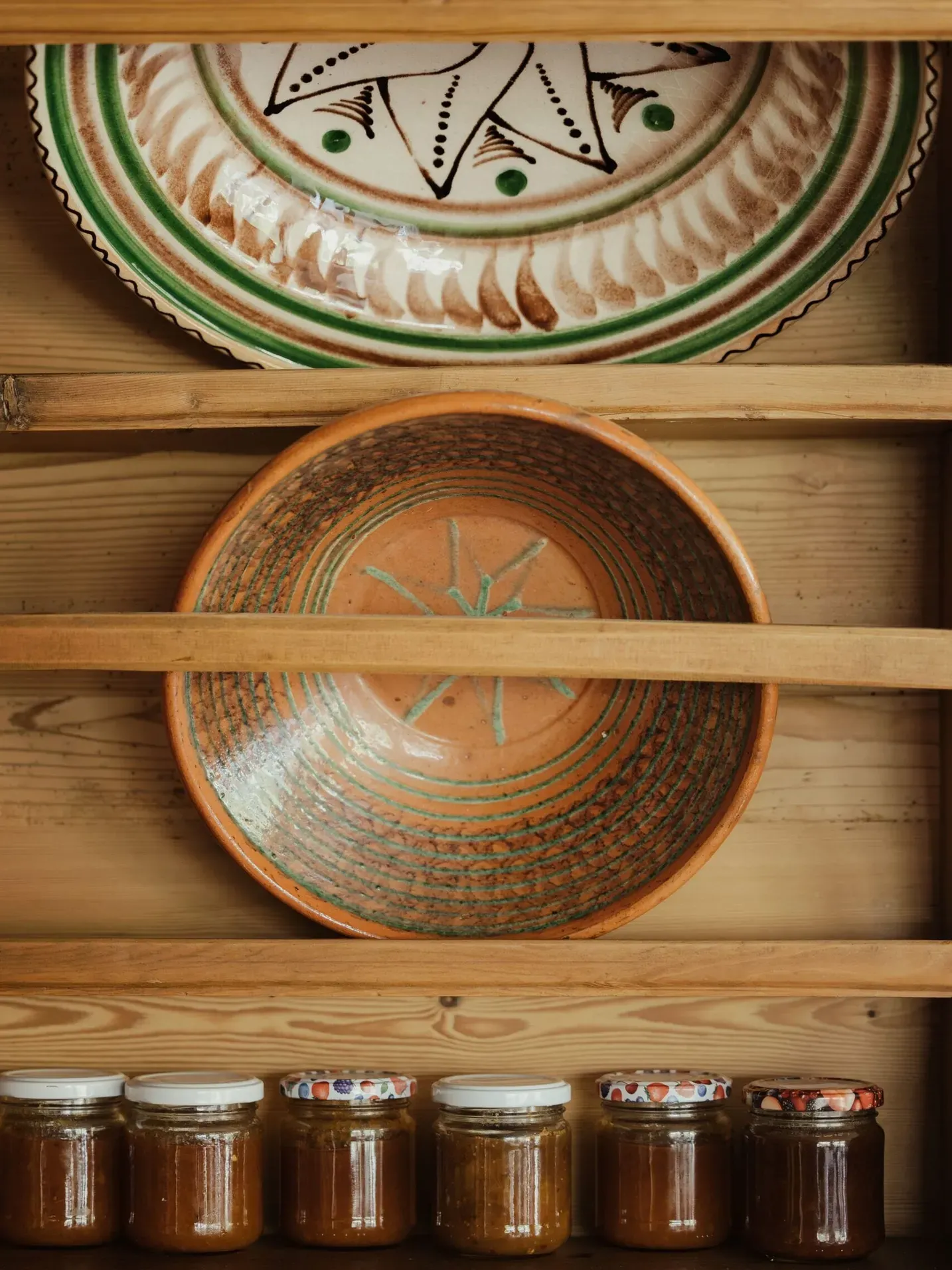 Mensole in legno con piatti decorativi e barattoli: in alto un grande piatto verde, al centro un piatto arancione, in basso vasetti di marmellata.