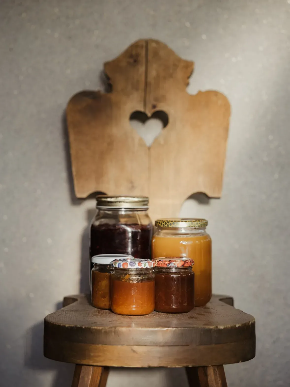 Barattoli di marmellata e miele su uno sgabello di legno, sfondo rustico con una tavola incisa a cuore; atmosfera calda e domestica.