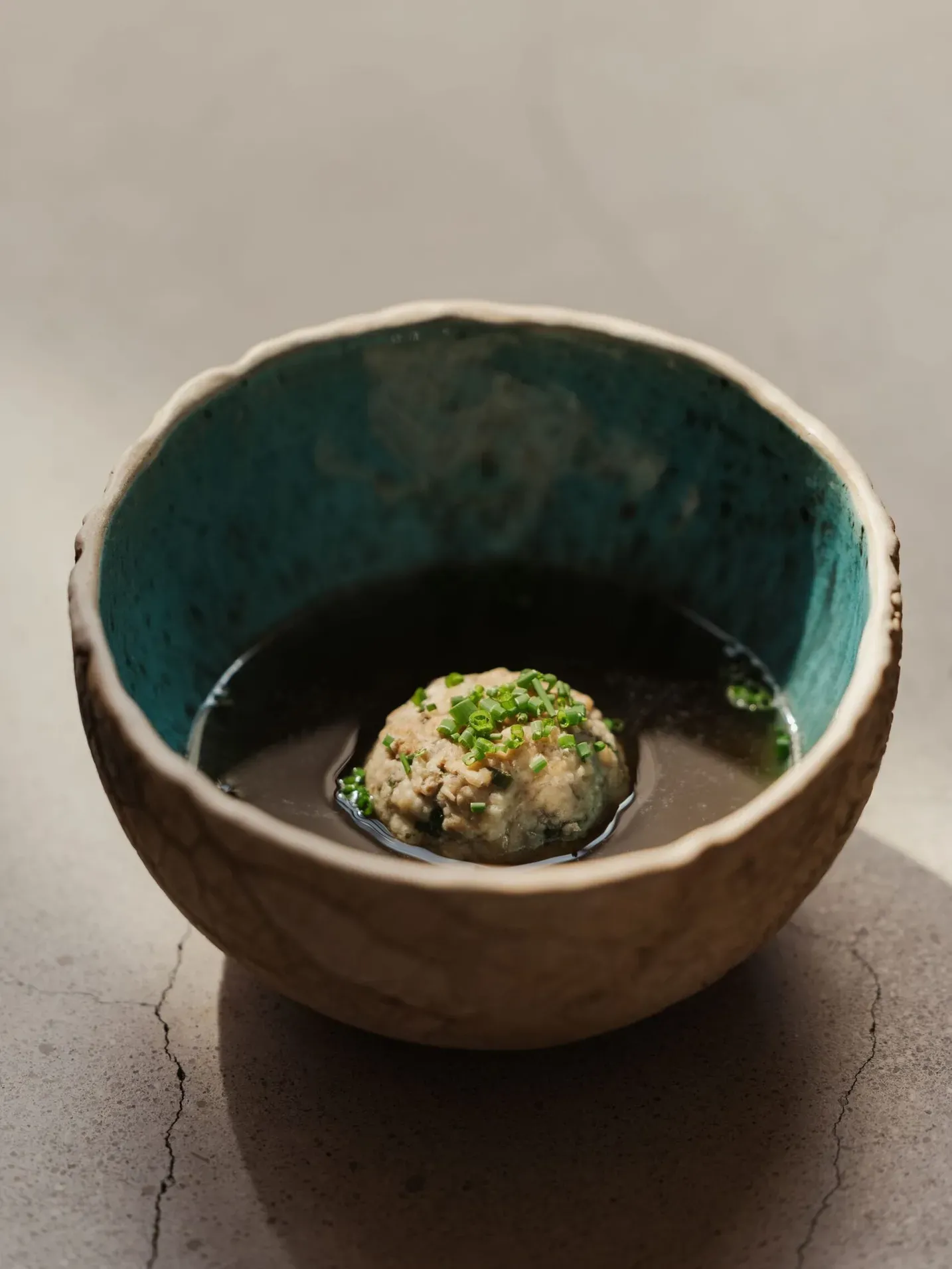Ciotola di ceramica grezza con brodo scuro e una polpetta al centro, guarnita con erba cipollina; interno azzurro-turchese.
