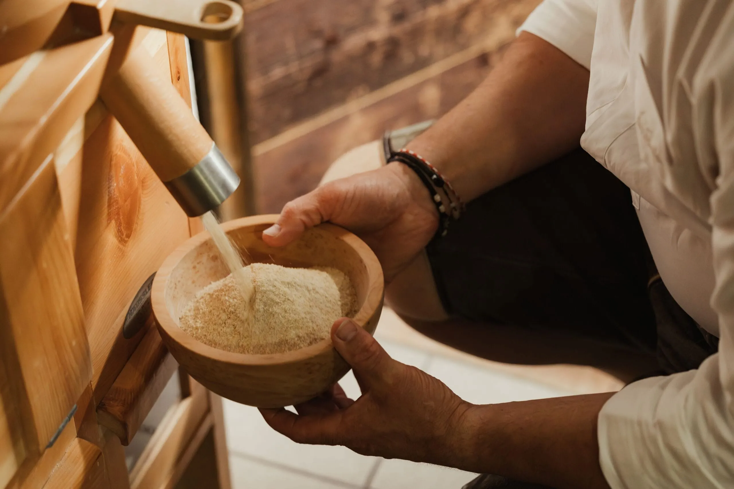 Person in a white shirt pours water from a metal spout into a wooden bowl of floury grains, with wooden milling equipment visible in the background.