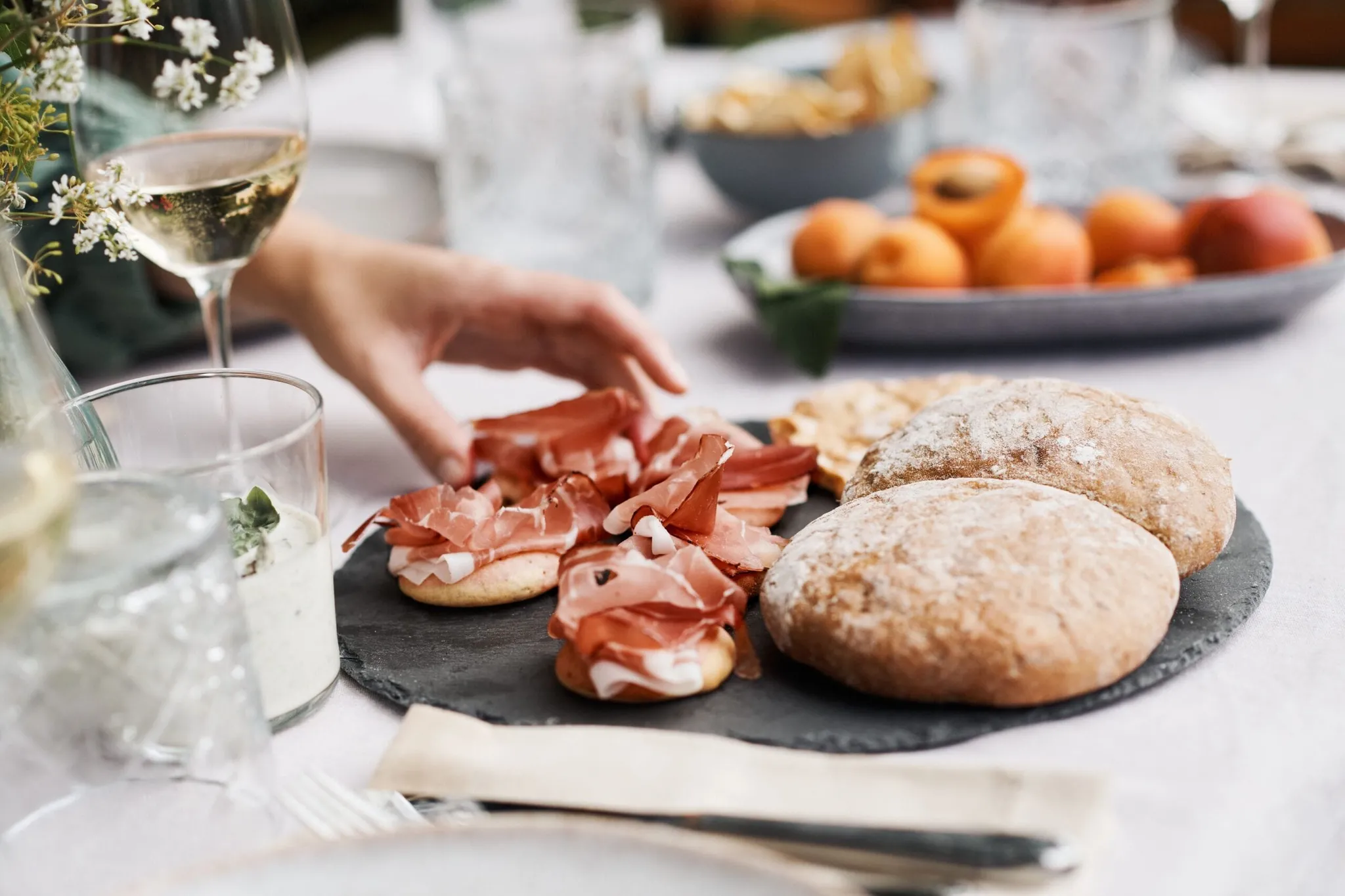 Slate board with crusty rolls and prosciutto crostini; a hand arranges meat, while a wine glass and peaches sit nearby in the background.
