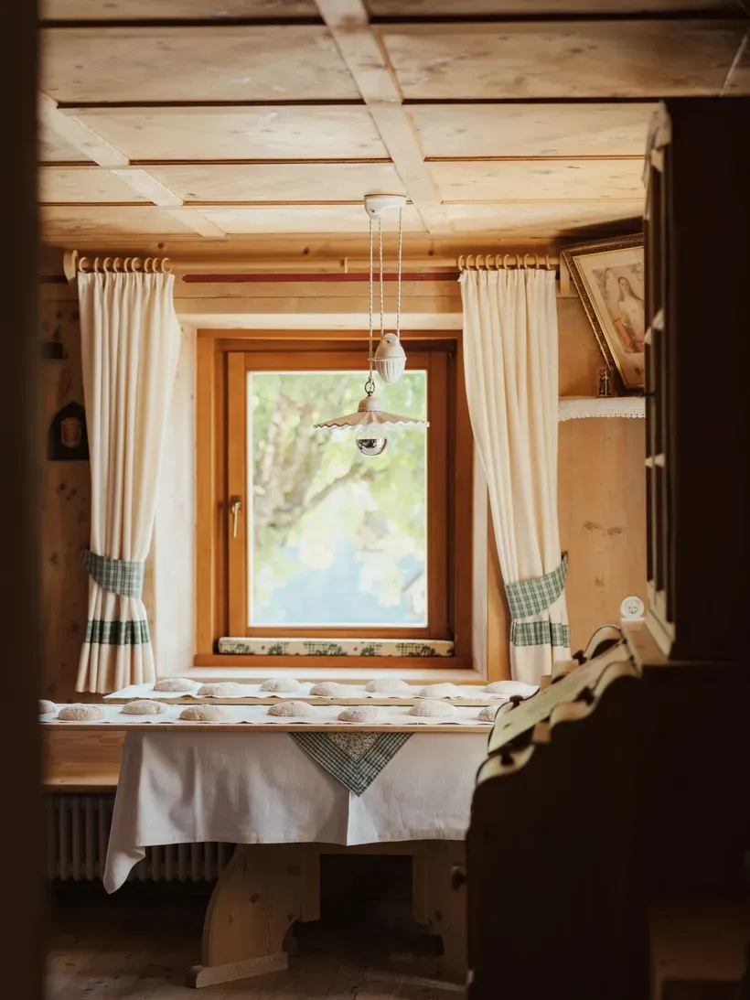 Cozy rustic room with a wooden ceiling and a window framed by white curtains; a long table with a white cloth and many small bread loaves, warm light.