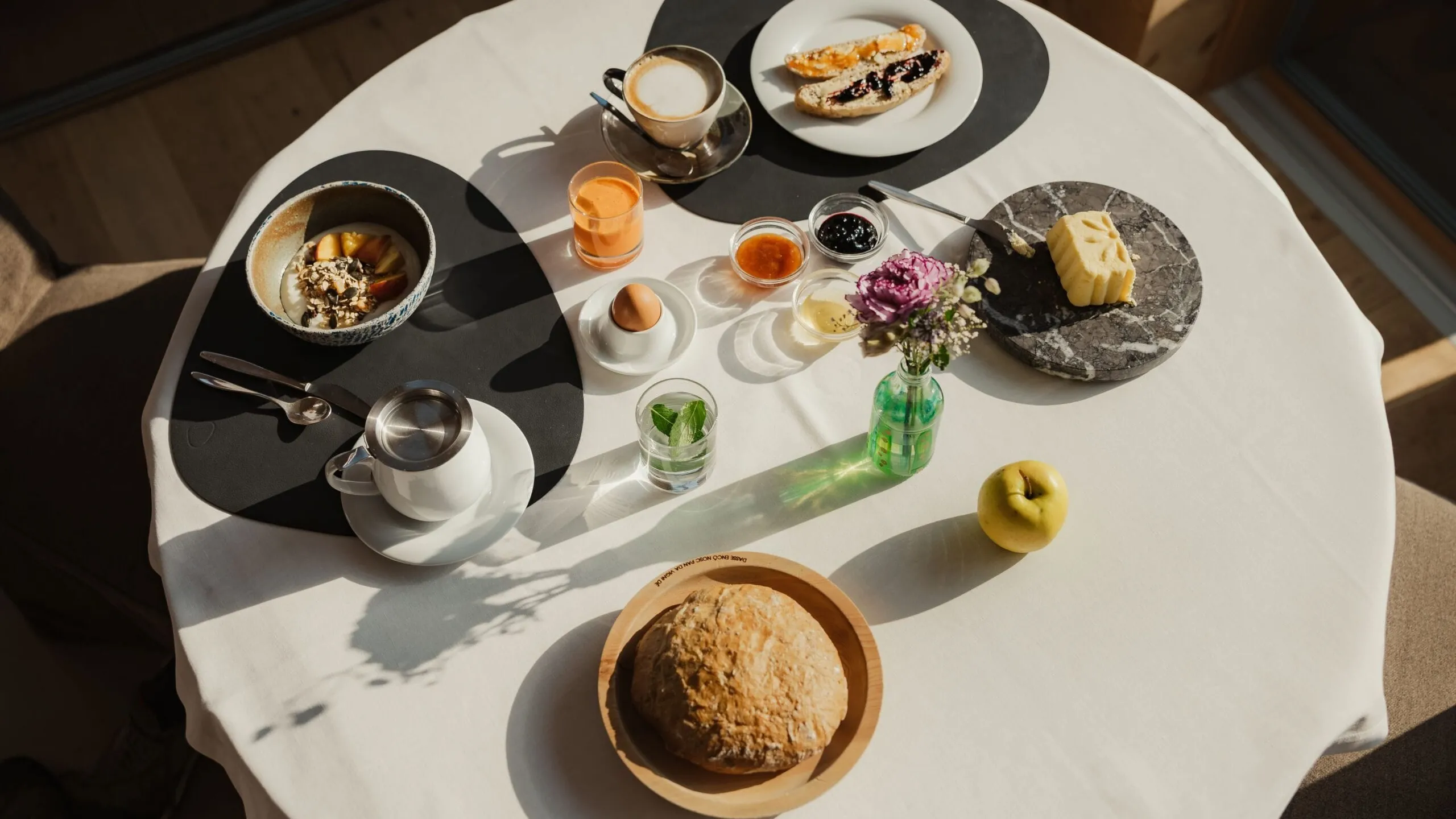Overhead view of a round white table set for breakfast: bread, apple, cheese on marble board, pastries, jams, coffee, an egg cup, a glass, and a small bouquet of flowers.