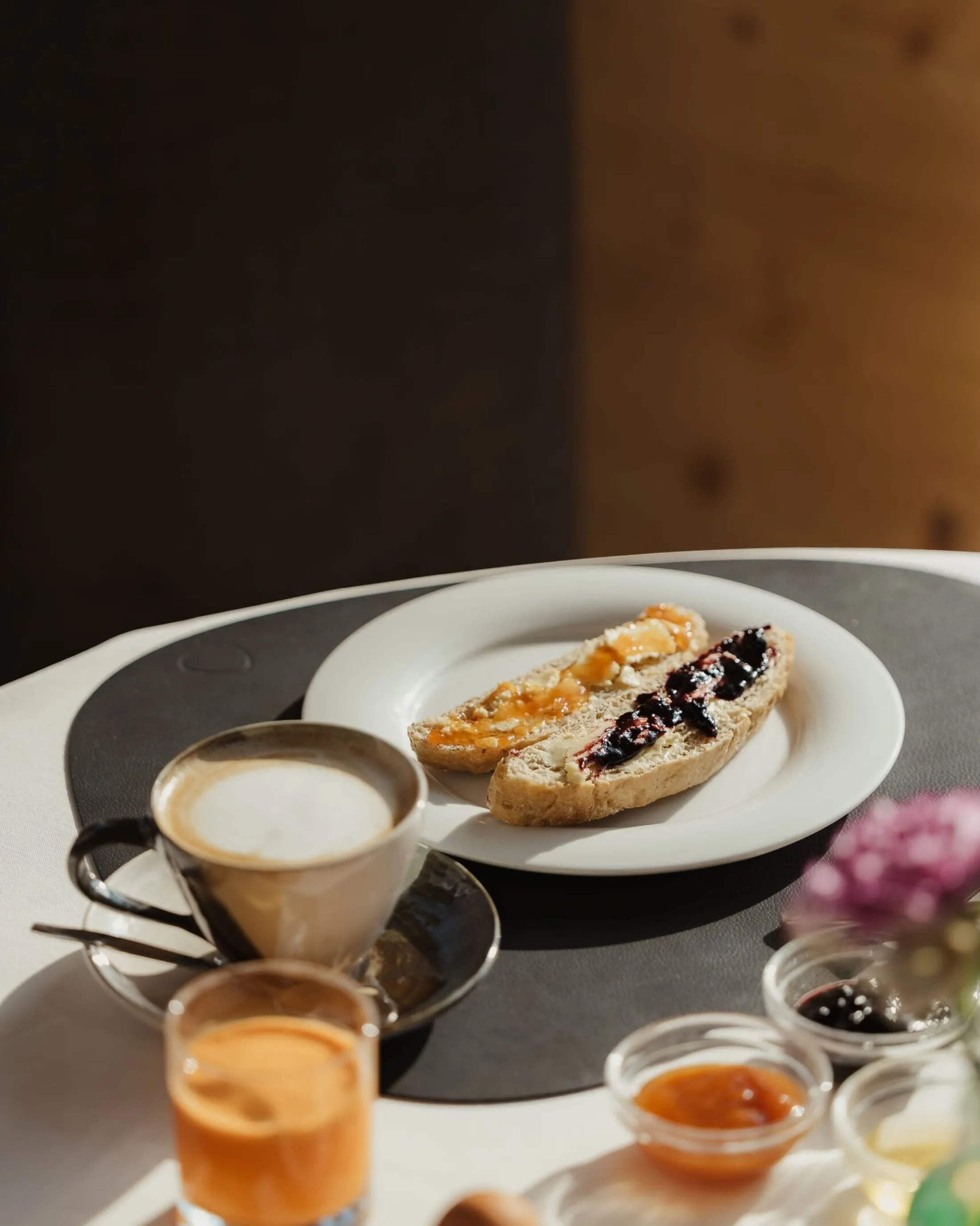 Colazione in hotel: pane con marmellata, cappuccino, succo d’arancia e piccoli vasetti di marmellata, con sfondo floreale sfocato.