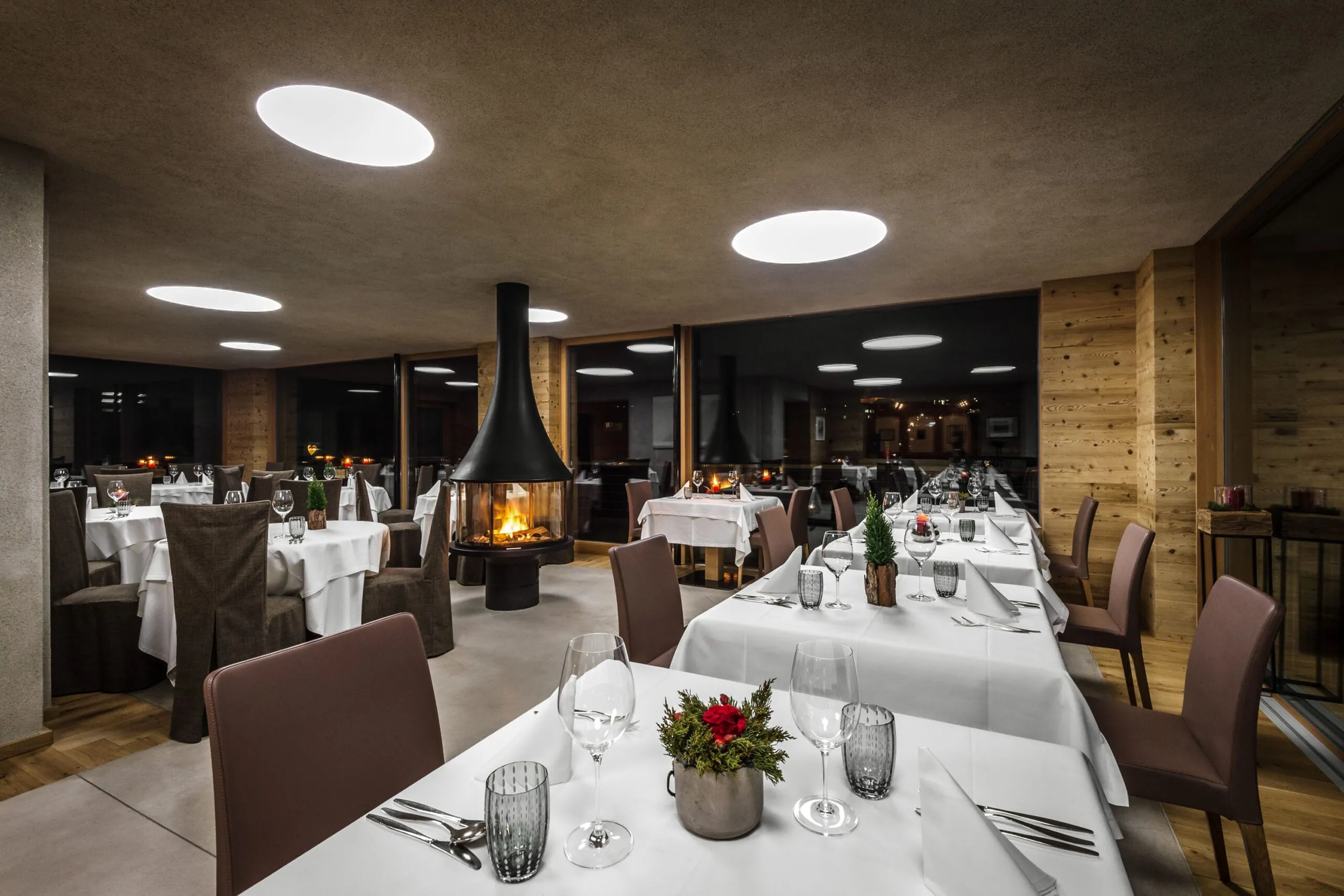 Hotel Gran Ander dining room in Badia, Alta Badia: white-tablecloth tables, brown chairs, a central hanging fireplace, wood paneling, round lights.