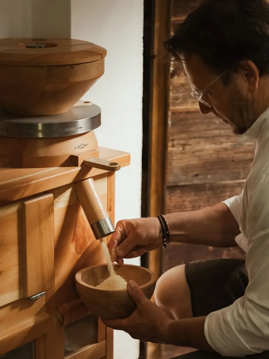 A person wearing a white shirt and glasses operates a wooden grain mill, pouring flour into a carved wooden bowl in a warm, rustic interior.